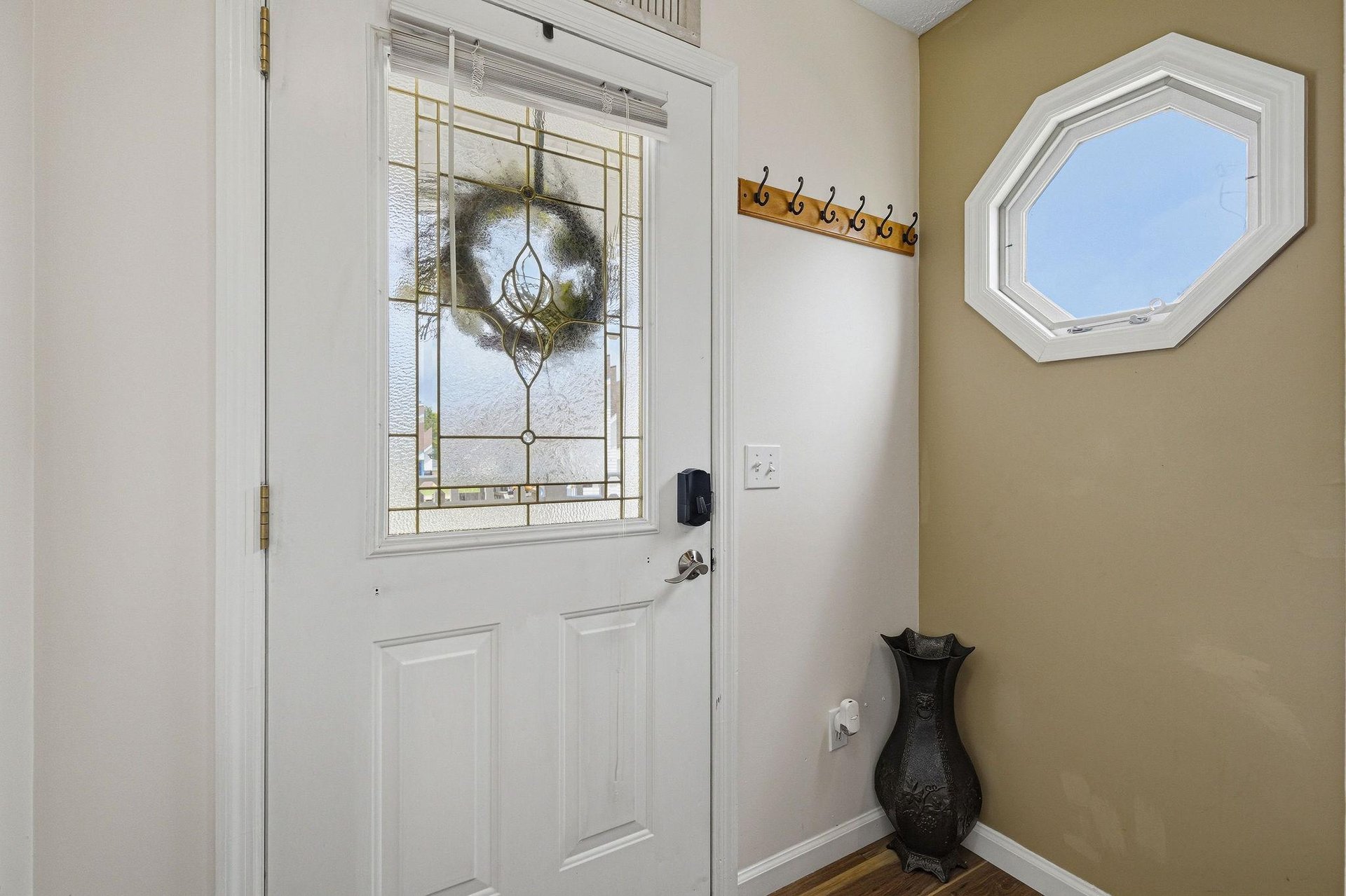 A welcoming entrance greets arrivals, featuring decorative stained glass, a charming wreath, and practical hooks for jackets and bags. Natural light streams through the unique octagonal window above.