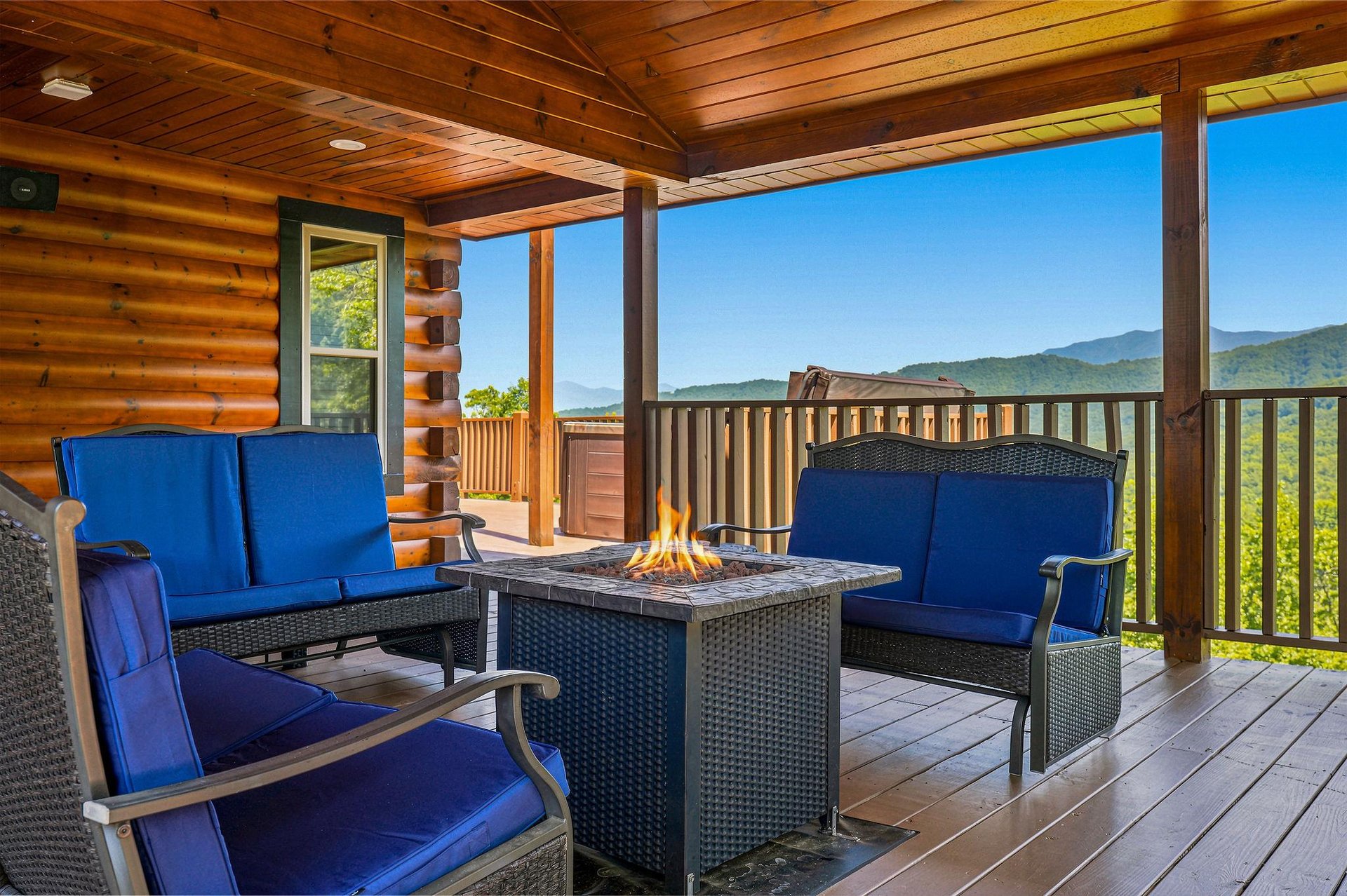 Relax on your private covered deck with comfortable seating around a fire table, taking in the breathtaking mountain views that stretch across the horizon under a clear blue sky.