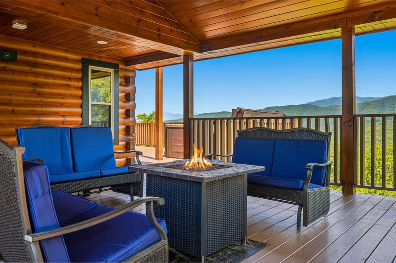 Relax on your private covered deck with comfortable seating around a fire table, taking in the breathtaking mountain views that stretch across the horizon under a clear blue sky.