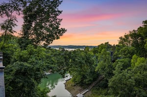 The propertys natural surroundings showcase a peaceful lake cove framed by lush trees, with vibrant sunset hues painting the sky over distant hills—a scenic backdrop to this waterfront retreat.