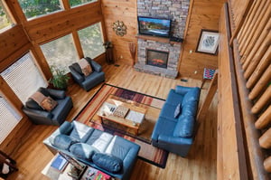 Soaring windows frame forest views in this cozy log cabin living room. Sink into plush seating by the stone fireplace and let the natural light and warmth embrace your mountain retreat.