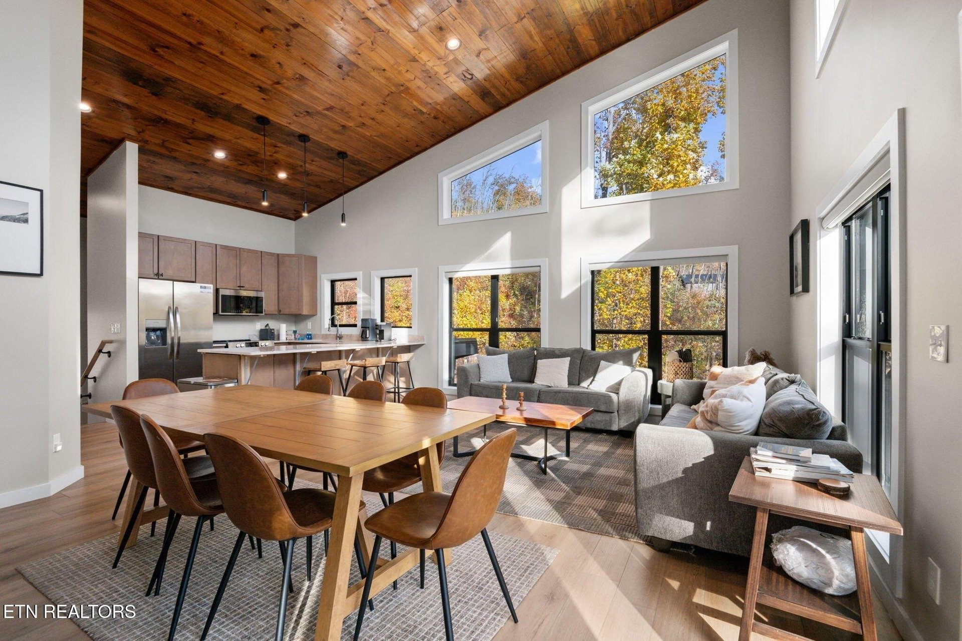 Step into an open, airy retreat where soaring ceilings with rich wood planks draw your eyes upward. Sunlight pours through floortoceiling windows, framing vibrant autumn views that make every moment