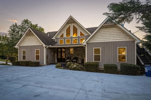 The property showcases a distinctive architectural design with its peaked gables and warm lighting, framed by mature trees and manicured landscaping. A welcoming entrance features stone accents and a 