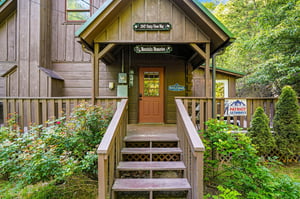 Charming mountain cabin entrance with rustic wood siding and welcoming front steps surrounded by lush greenery.