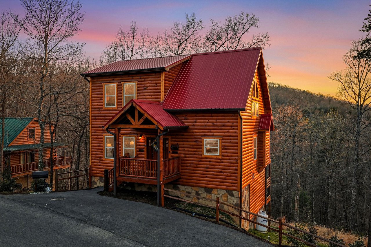 Mountain cabin with log exterior and red metal roof nestled among winter trees, featuring covered porch entrance and scenic wooded surroundings.