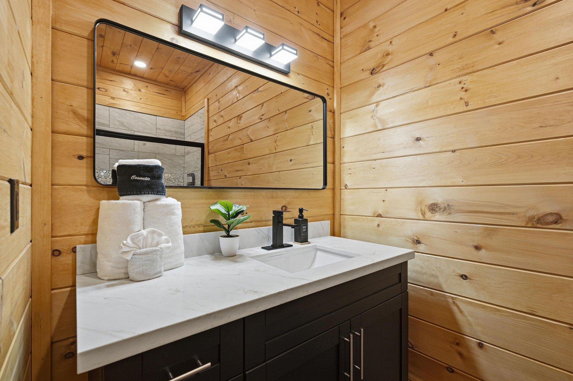 Freshen up in your beautifully appointed bathroom featuring warm wood walls, modern vanity, and thoughtful touches like fluffy towels and live greenery.