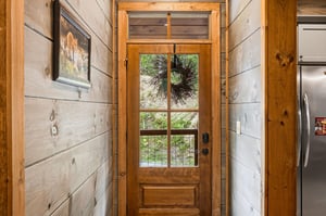 A welcoming entryway framed by warm shiplap walls, featuring a glasspaneled door adorned with seasonal greenery, opening to views of natural surroundings beyond.