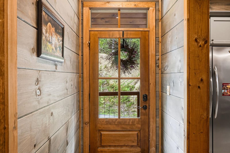 A welcoming entryway framed by warm shiplap walls, featuring a glasspaneled door adorned with seasonal greenery, opening to views of natural surroundings beyond.