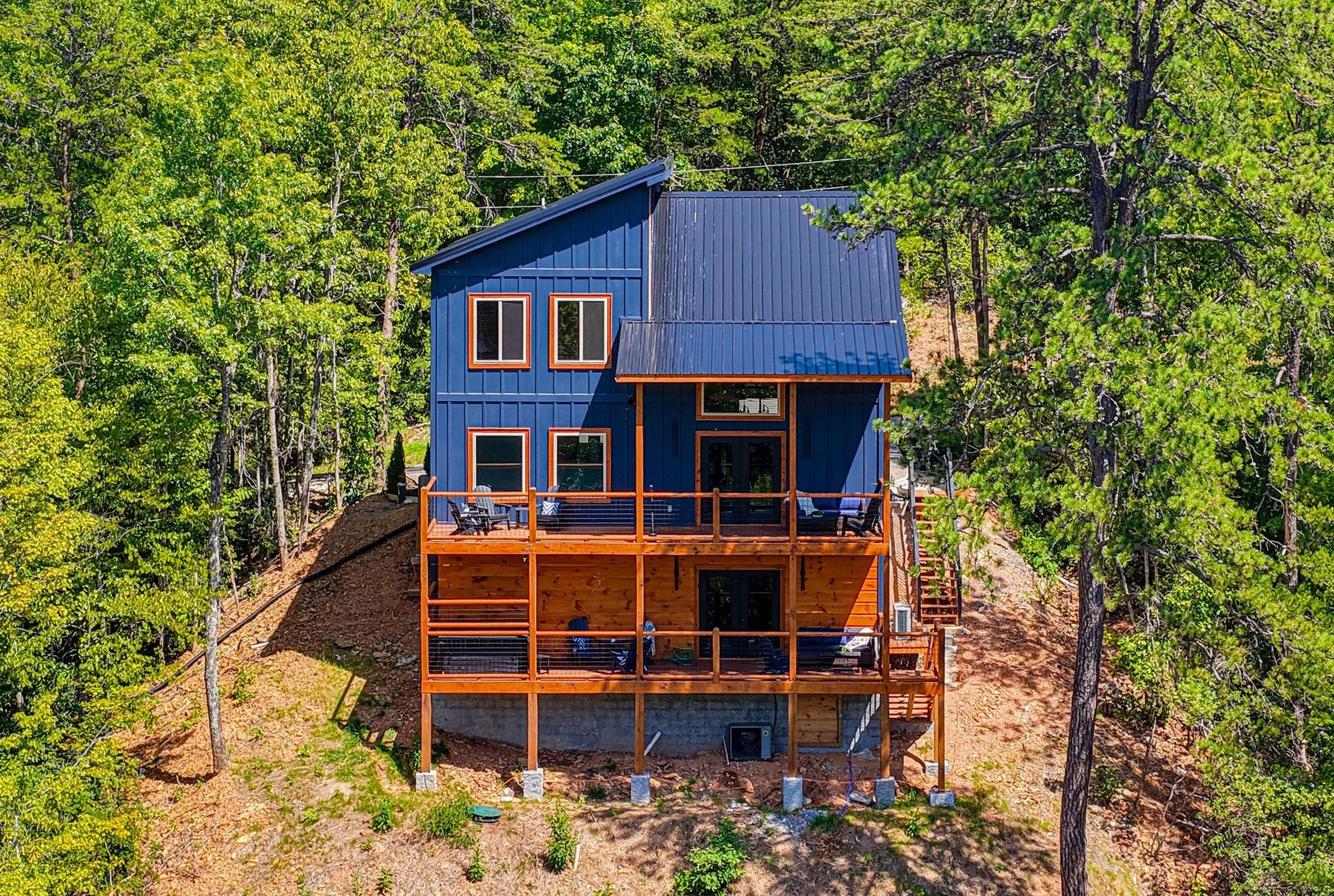 A modern blue cabin nestled among vibrant green trees, featuring expansive wood decks and generous windows capturing the natural beauty surrounding the property.