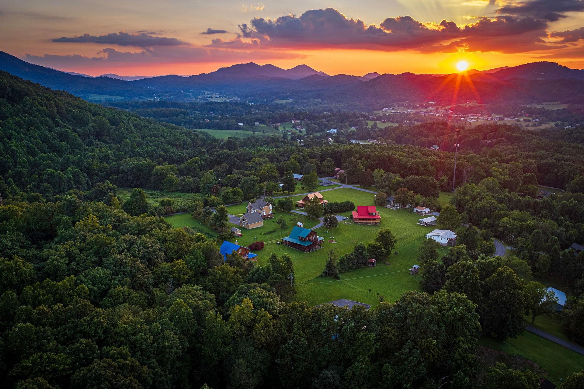 A breathtaking aerial view captures the property nestled in a lush valley, surrounded by rolling hills and majestic mountains, as the sun sets in a blaze of golden light.