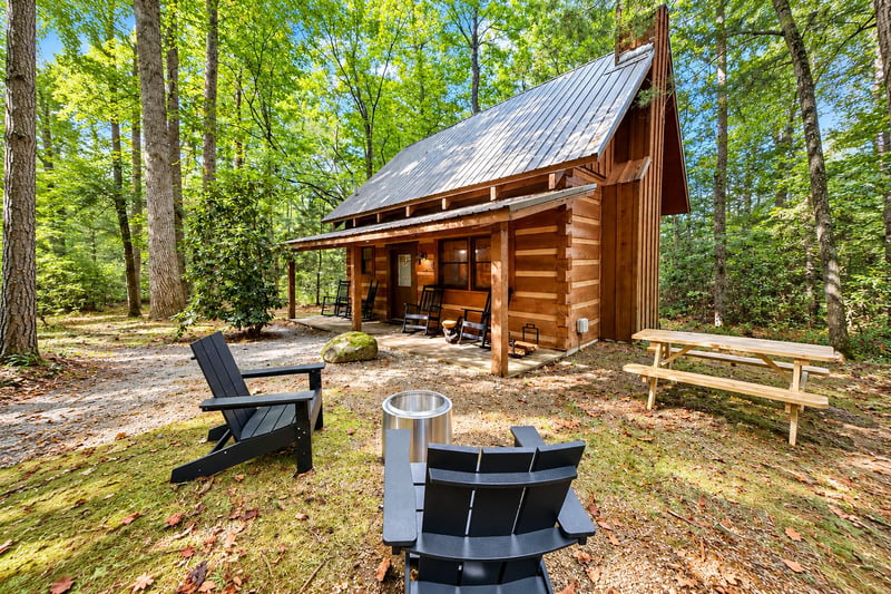 A charming log cabin nestled in a lush forest offers peaceful woodland surroundings. Relax by the fire pit or unwind on the covered porch with rocking chairs just steps away.