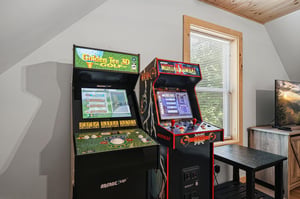 Your private game room awaits with classic arcade machines ready for friendly competition, framed by natural light and cozy woodbeam ceilings overhead.