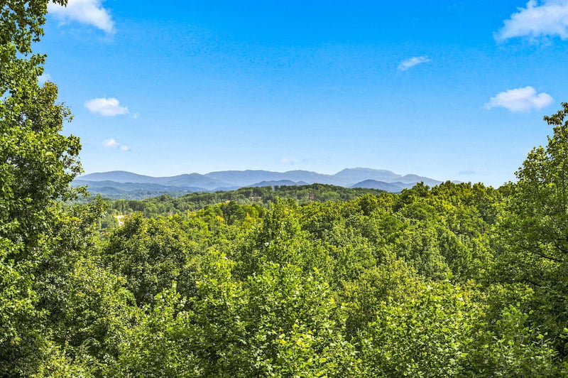 A property nestled amid lush wooded landscapes with majestic mountain ranges framing the horizon under a clear blue sky.