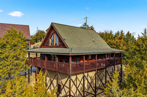 Elevated cabin nestled among towering trees, featuring wraparound deck and mountain architecture in a peaceful forest setting.