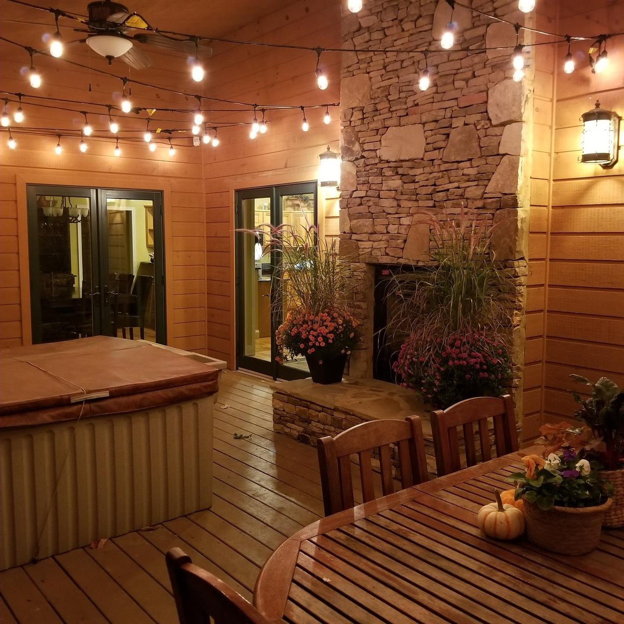 Your private covered patio glows with string lights at dusk, featuring a welcoming hot tub, outdoor dining table, and a rustic stone fireplace surrounded by seasonal blooms.