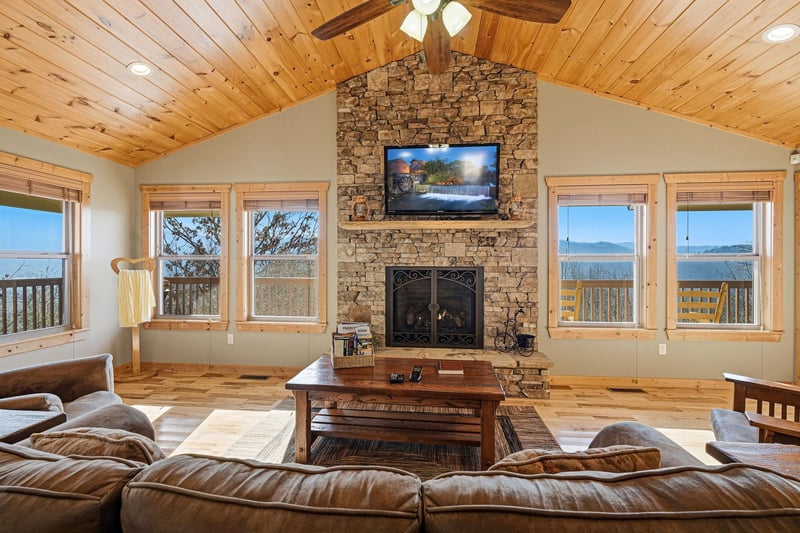Unwind in this cozy living room where rustic wood meets stone fireplace warmth, while panoramic mountain views stretch beyond every window.