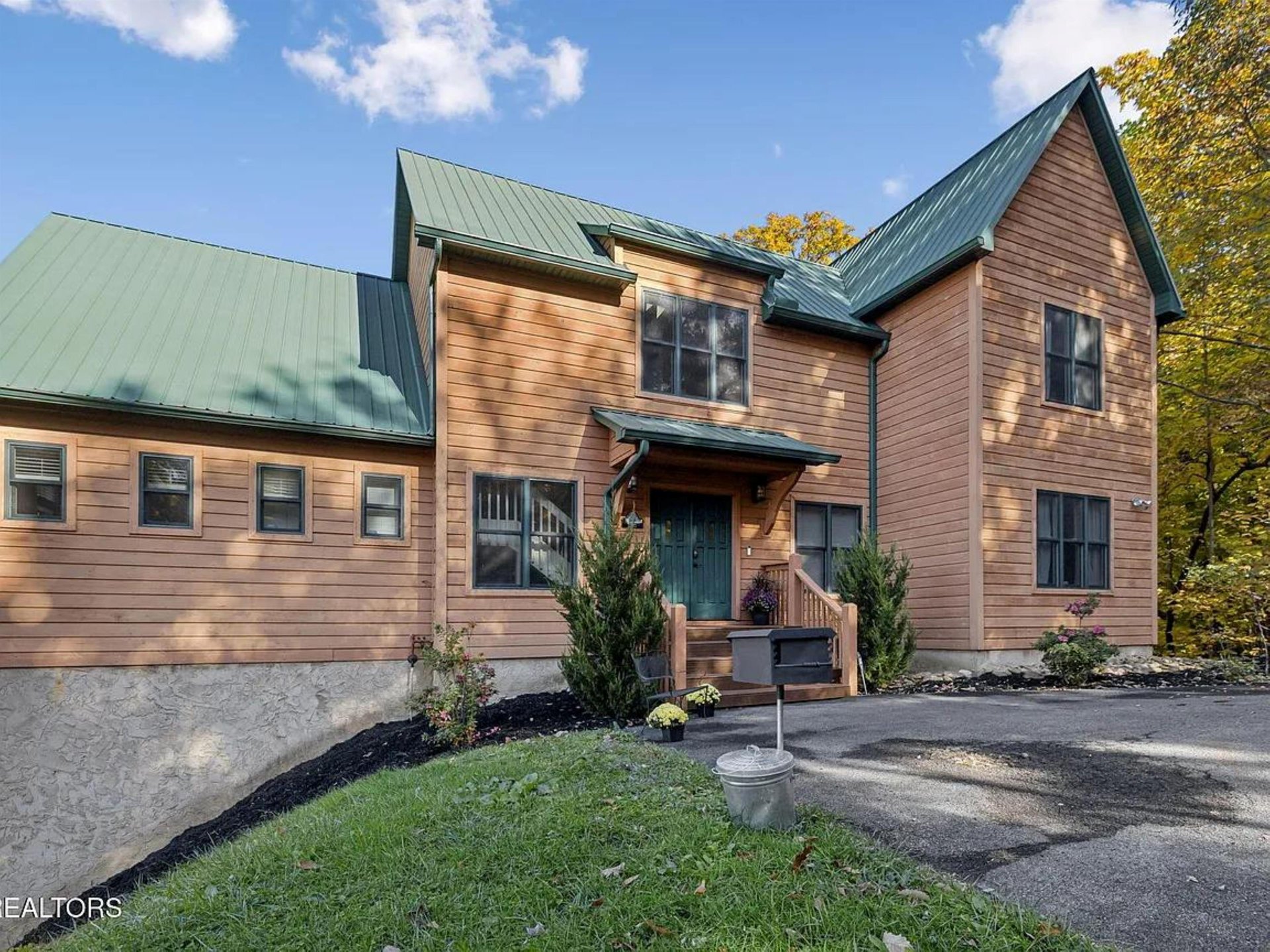 Charming mountain retreat featuring warm wood siding and green metal roofing, surrounded by autumn foliage. The property offers convenient parking and a welcoming front entrance with seasonal landscap