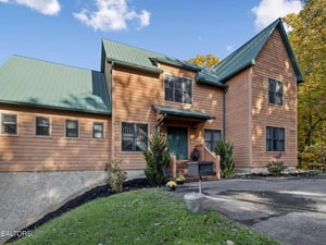 Charming mountain retreat featuring warm wood siding and green metal roofing, surrounded by autumn foliage. The property offers convenient parking and a welcoming front entrance with seasonal landscap