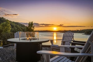 Sunset over calm water creates a stunning backdrop for this propertys outdoor gathering area, featuring a fire pit table and Adirondack chairs overlooking the scenic waterfront.