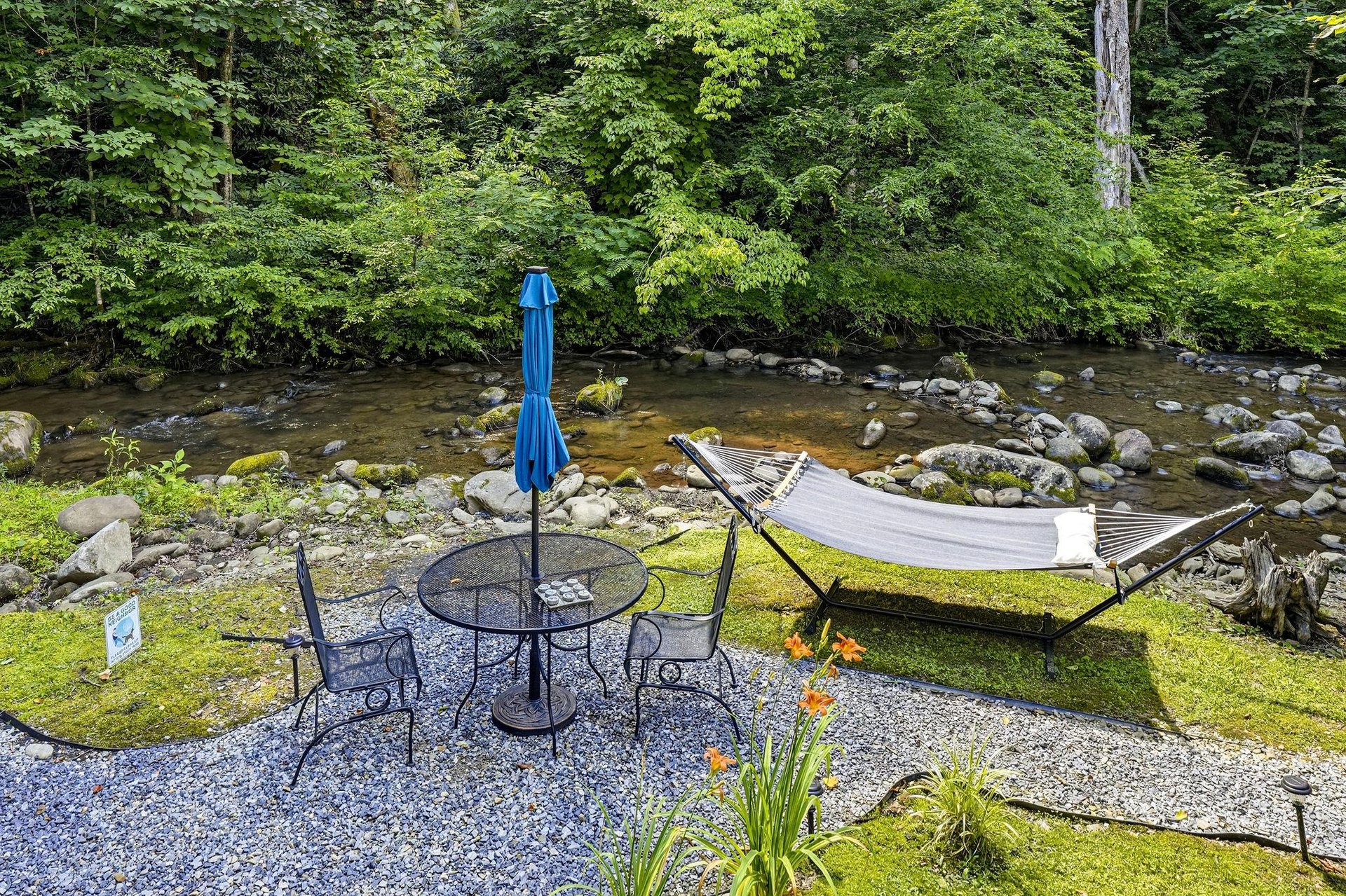 A tranquil riverside retreat where a gentle stream flows past lush forest banks, offering a peaceful patio with seating and a hammock for ultimate relaxation.