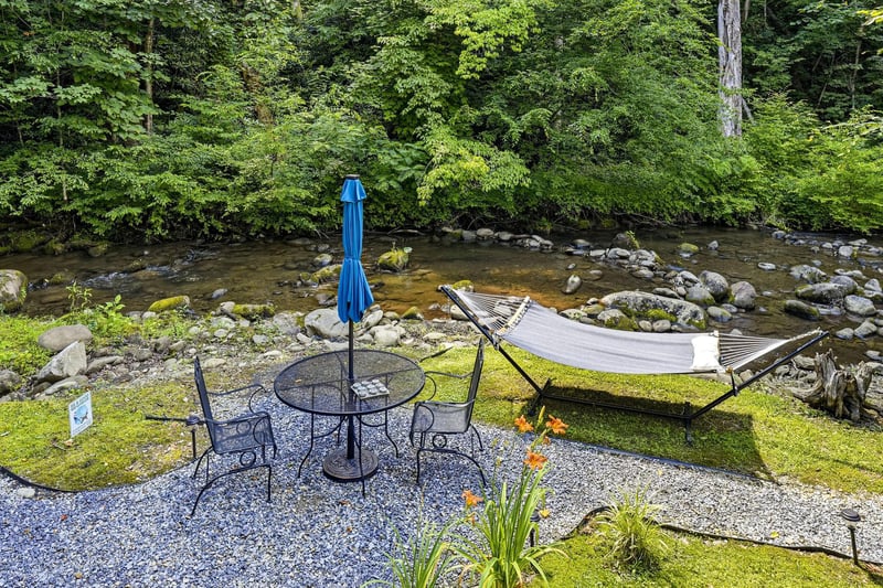 A tranquil riverside retreat where a gentle stream flows past lush forest banks, offering a peaceful patio with seating and a hammock for ultimate relaxation.