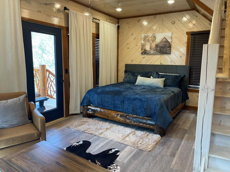 Your cozy bedroom features rustic wood walls and opens to a private balcony where morning light filters through tall trees.