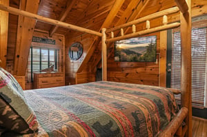 Your rustic retreat features a handcrafted log bed beneath soaring woodbeam ceilings, with a flatscreen TV displaying mountain views to enhance your cozy cabin experience.