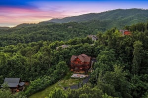 Aerial view of a spacious mountain lodge nestled among lush forested hills, capturing the serene natural landscape and scenic surroundings at sunset.