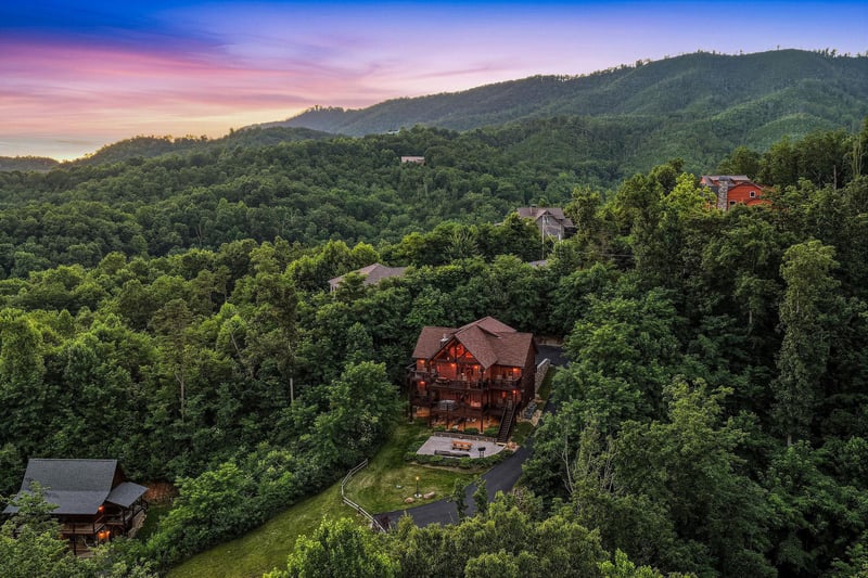 Aerial view of a spacious mountain lodge nestled among lush forested hills, capturing the serene natural landscape and scenic surroundings at sunset.