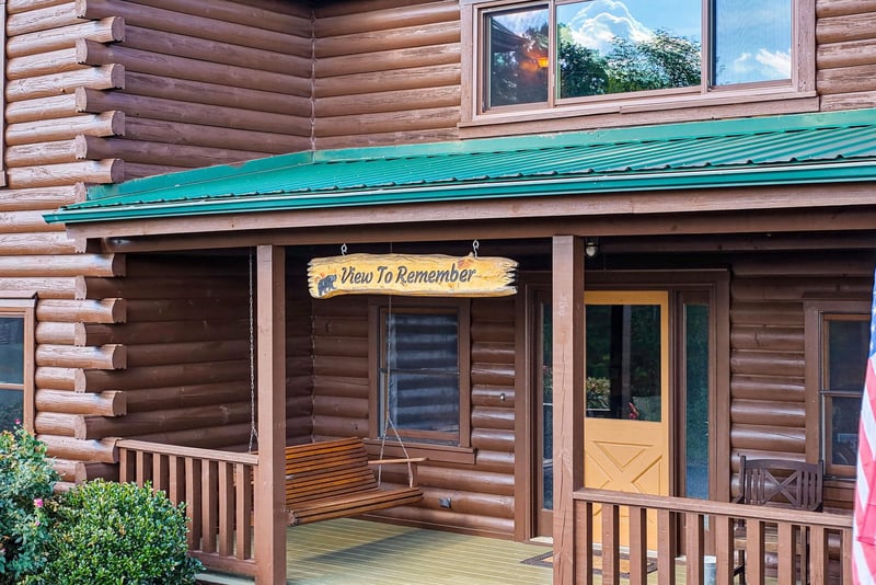 Charming log cabin with a welcoming covered porch, rustic swing, and green deck. The entrance opens to genuine mountain views, blending traditional log construction with natural surroundings.