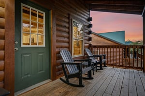 Rustic log cabin with green entrance and covered deck overlooking forested mountains, captured during the warm glow of sunset.