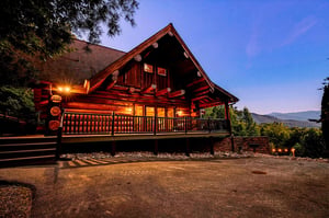 A beautiful log cabin property set among natural surroundings with mountain vistas in the distance, captured at twilight with warm lighting highlighting the handcrafted timber construction.