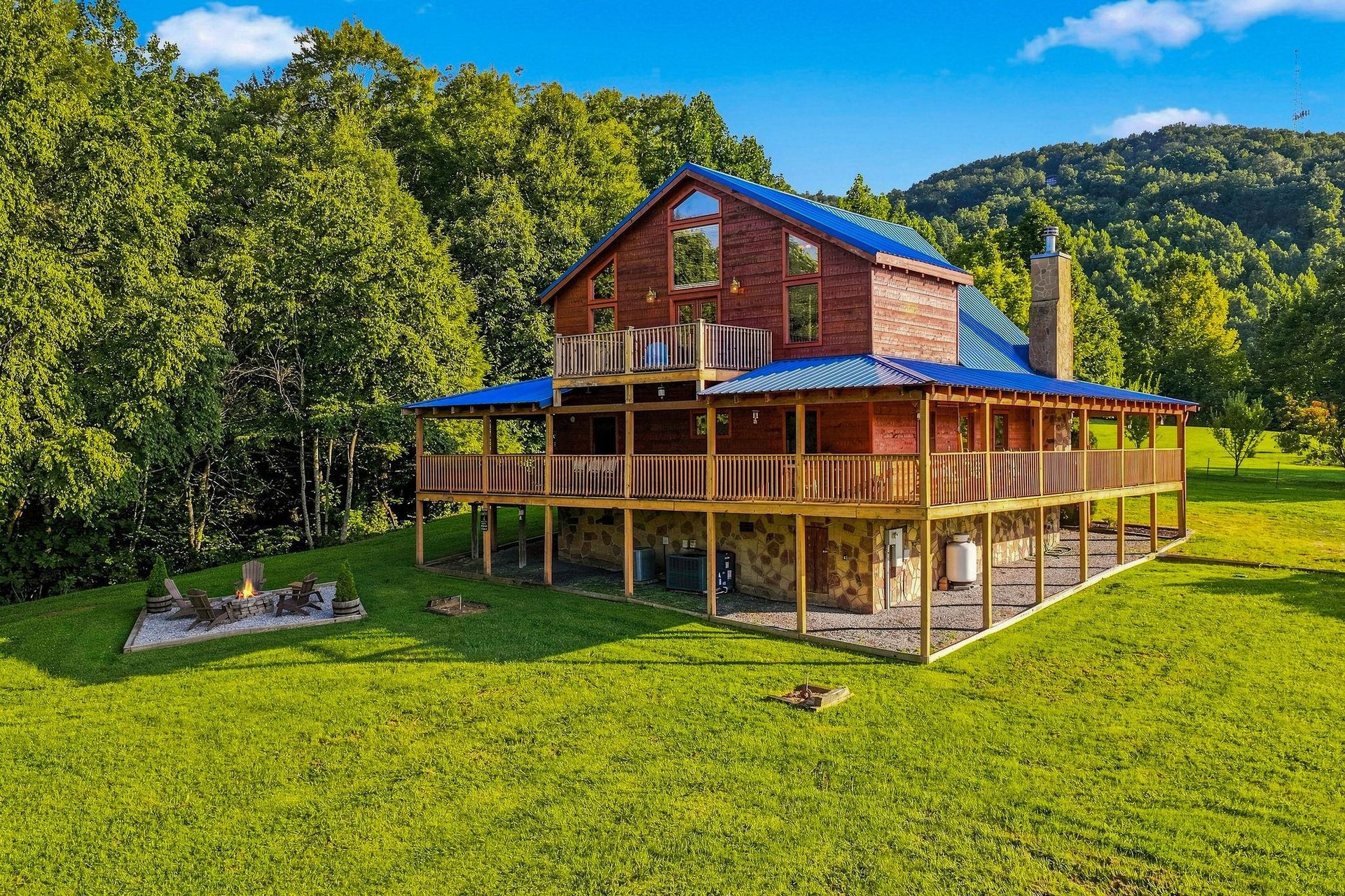 A distinctive mountain cabin with bright blue metal roof nestled against forested hillsides, featuring wraparound porches and expansive windows overlooking lush green lawns and fire pit area.