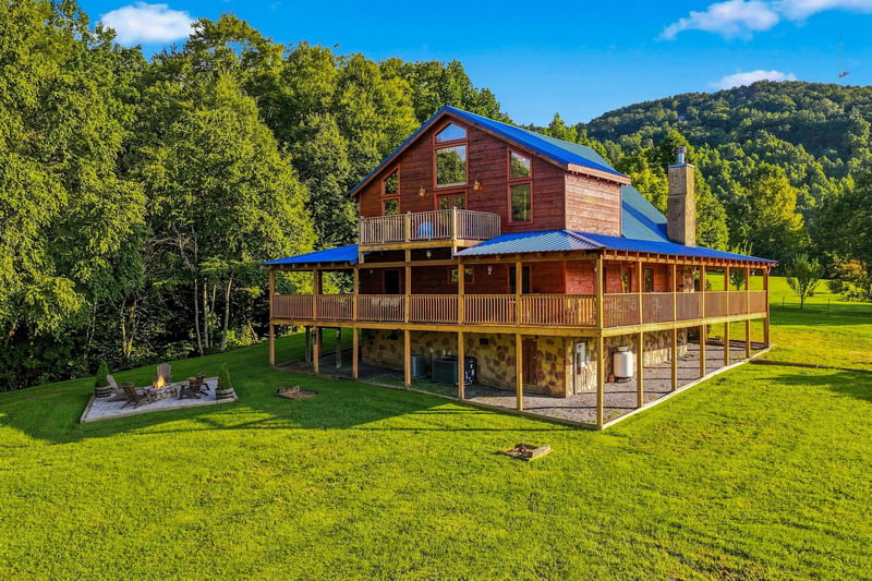 A distinctive mountain cabin with bright blue metal roof nestled against forested hillsides, featuring wraparound porches and expansive windows overlooking lush green lawns and fire pit area.