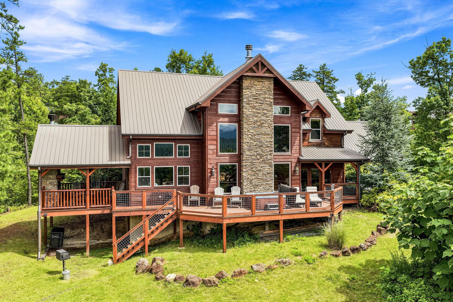 A striking cabin with natural wood siding, stone chimney, and expansive wraparound deck set on lush grounds surrounded by towering trees.