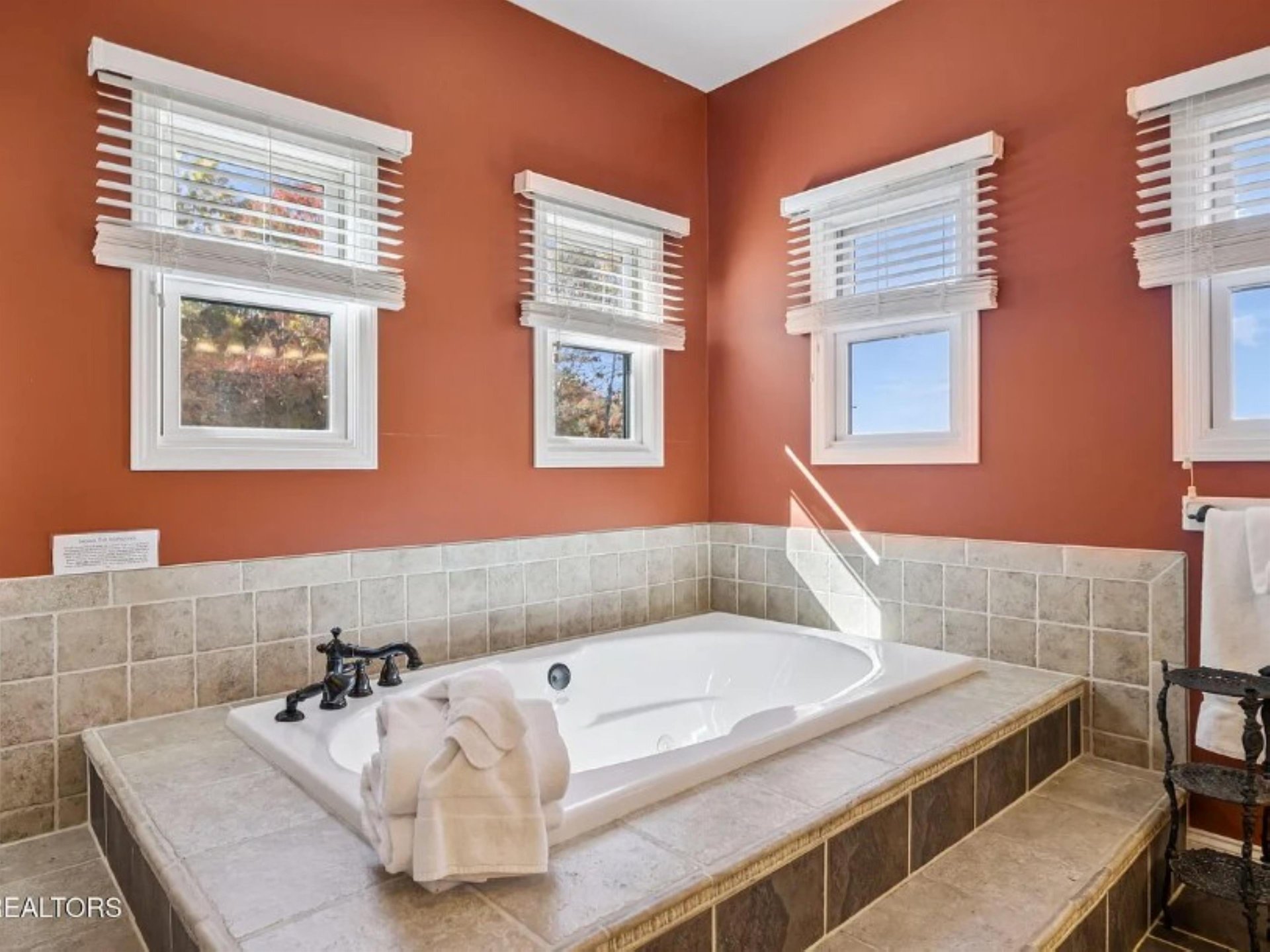 Picture yourself unwinding in this elegant soaking tub surrounded by warm terracotta walls and natural light streaming through four windows. The tiled surround and plush towels create your perfect spa