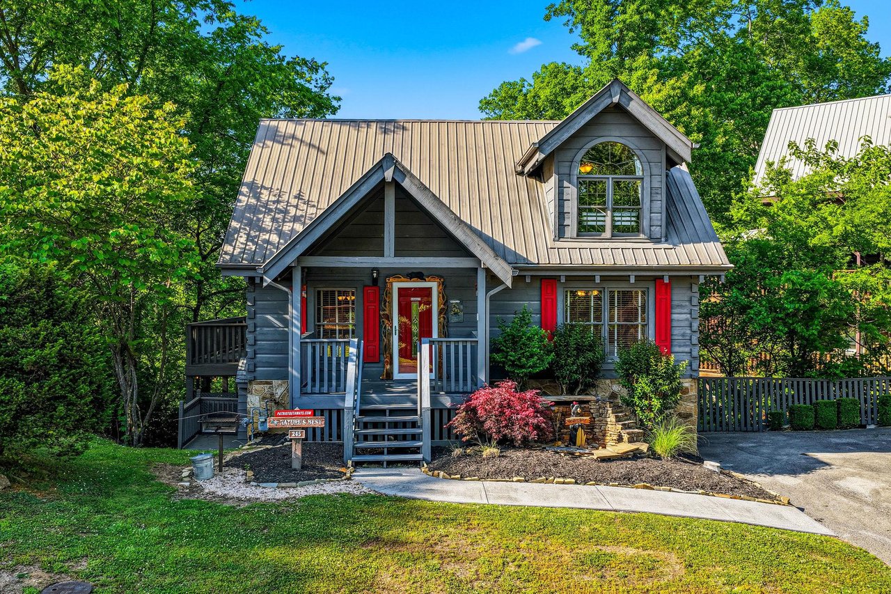 Charming mountain cabin with striking red shutters nestled among mature trees, featuring welcoming front porch and landscaped grounds.