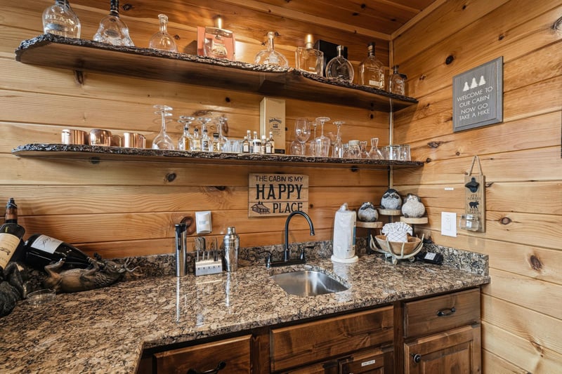 Your cabin retreat features a welcoming wet bar with granite countertops, rustic wooden shelving displaying glassware, and charming cabinthemed décor—perfect for unwinding with your favorite drink.