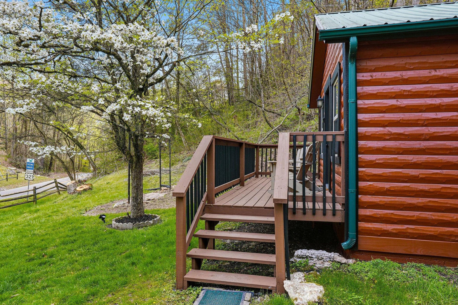 Rustic log cabin entrance with deck surrounded by blooming spring trees and peaceful countryside setting.