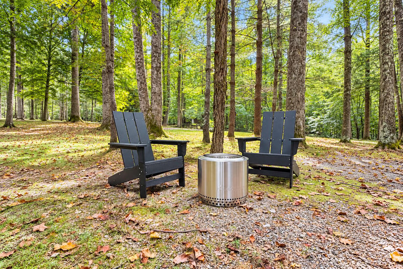 Nestled in a tranquil forest setting with towering trees overhead and seasonal foliage underfoot, your private outdoor fire pit area awaits with two comfortable Adirondack chairs for peaceful moments 
