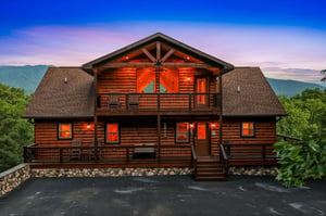 A handsome log cabin glows warmly against a twilight sky, nestled among forested mountains with spacious decks and scenic surroundings.