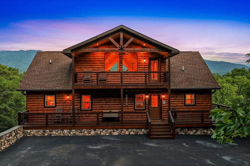 A handsome log cabin glows warmly against a twilight sky, nestled among forested mountains with spacious decks and scenic surroundings.