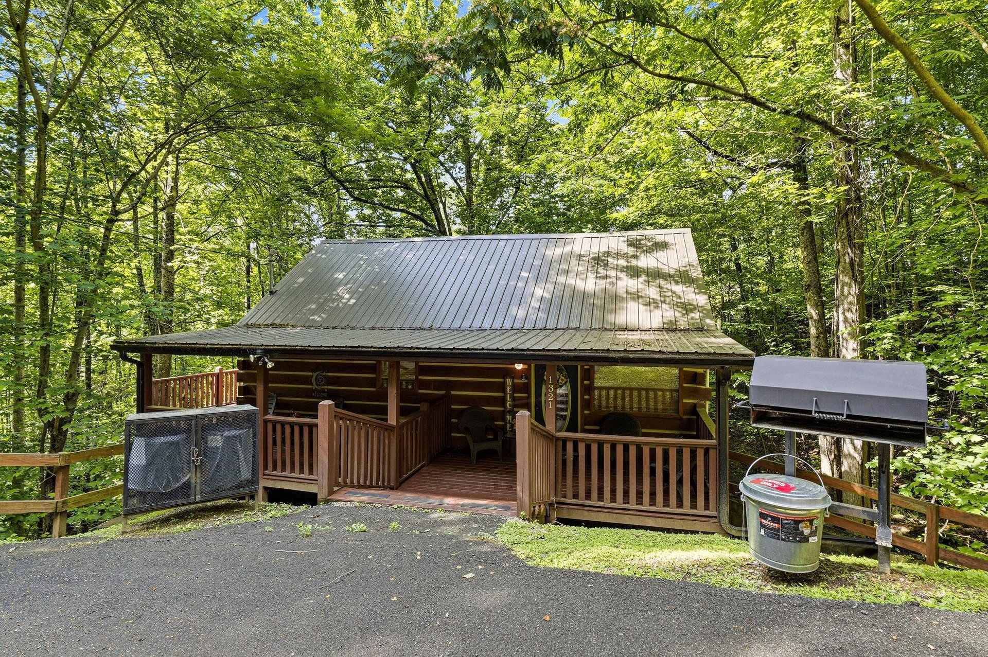 A charming log cabin surrounded by lush forest, featuring a welcoming front porch, convenient parking, and BBQ facilities for outdoor meals.