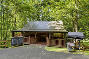 A charming log cabin surrounded by lush forest, featuring a welcoming front porch, convenient parking, and BBQ facilities for outdoor meals.