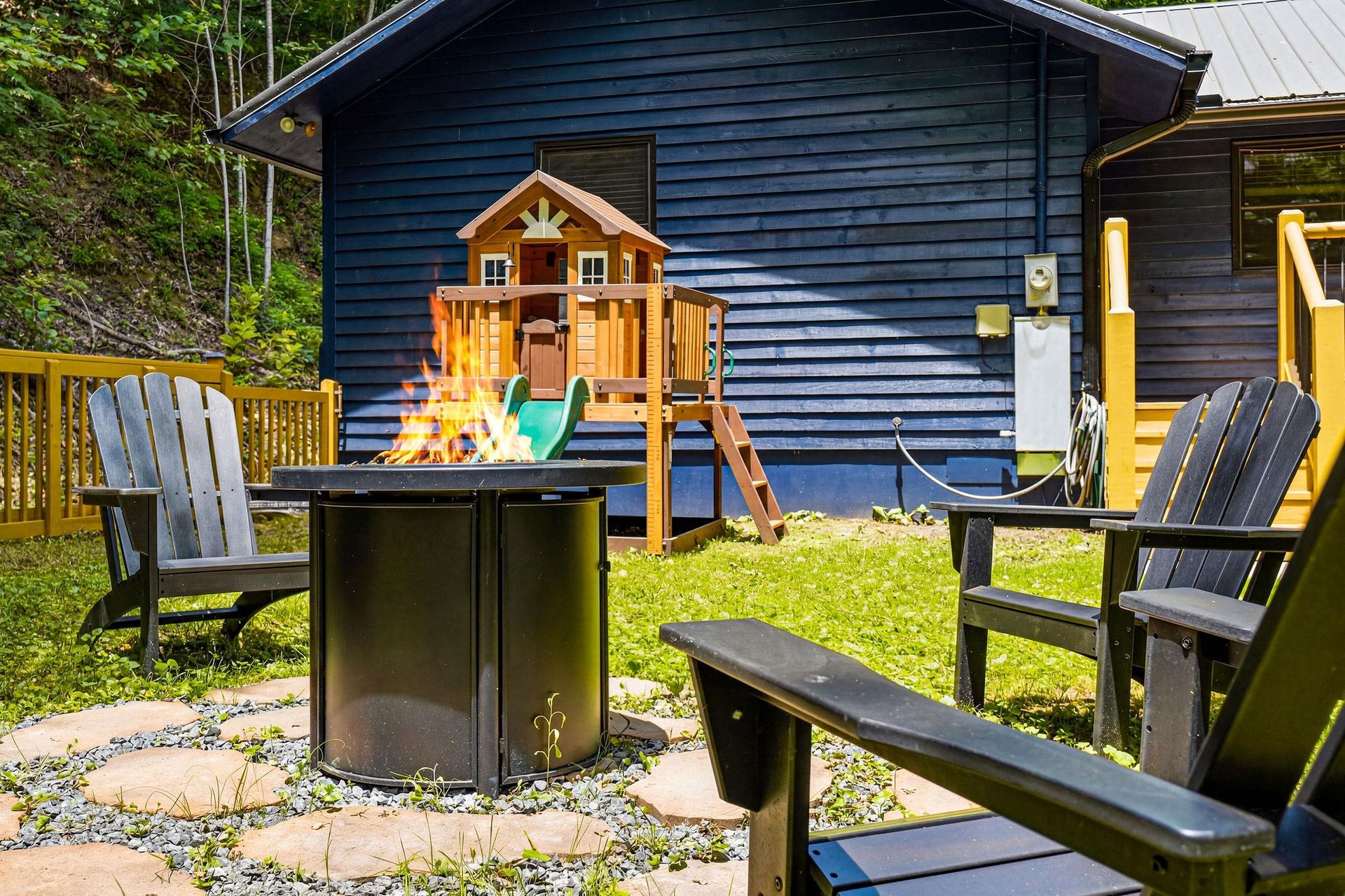 A welcoming backyard retreat features cozy outdoor seating by the fire pit, a playground for little ones, and lush greenery all around—perfect for family gatherings and peaceful summer days.