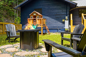 A welcoming backyard retreat features cozy outdoor seating by the fire pit, a playground for little ones, and lush greenery all around—perfect for family gatherings and peaceful summer days.