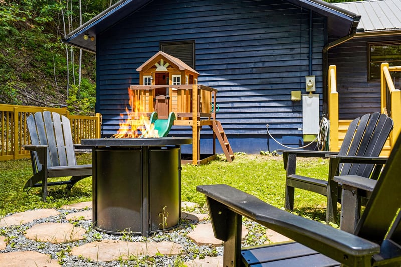 A welcoming backyard retreat features cozy outdoor seating by the fire pit, a playground for little ones, and lush greenery all around—perfect for family gatherings and peaceful summer days.