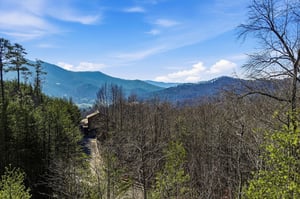 Panoramic mountain views stretch across rolling forested hills beneath a bright blue sky dotted with white clouds.