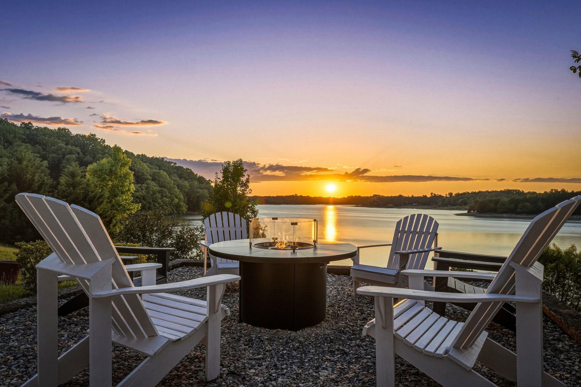 Lakeside fire pit with Adirondack seating captures golden sunset reflections across the tranquil water, surrounded by forested shores and peaceful evening skies.