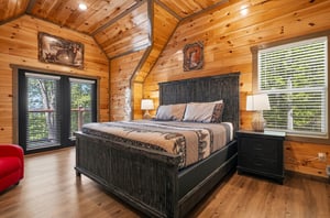 Unwind in your rustic bedroom featuring warm wood walls, cozy bedding, and peaceful forest views through large windows.